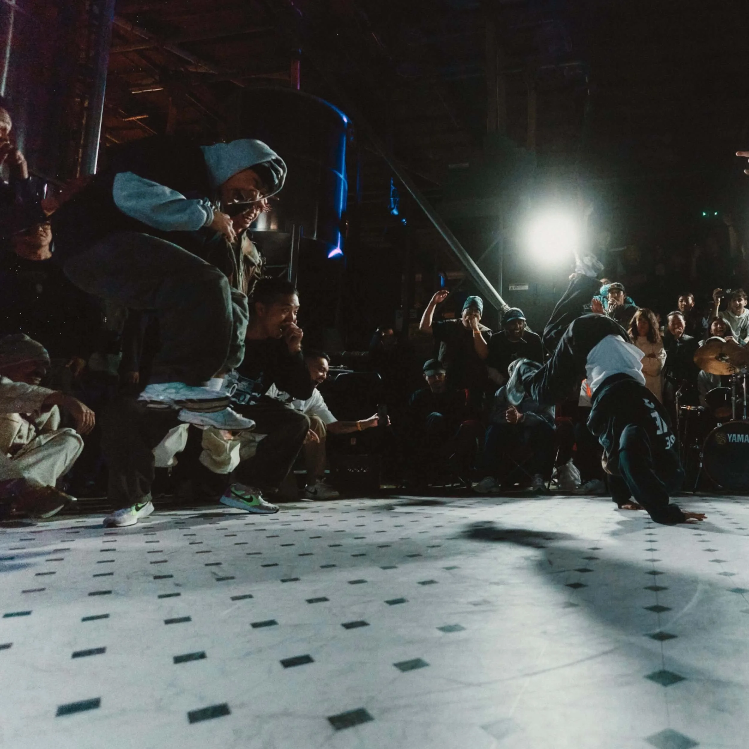 A b-boy frozen in a headspin on a checkered floor as a dim crowd watches — Elephant Graveyard.