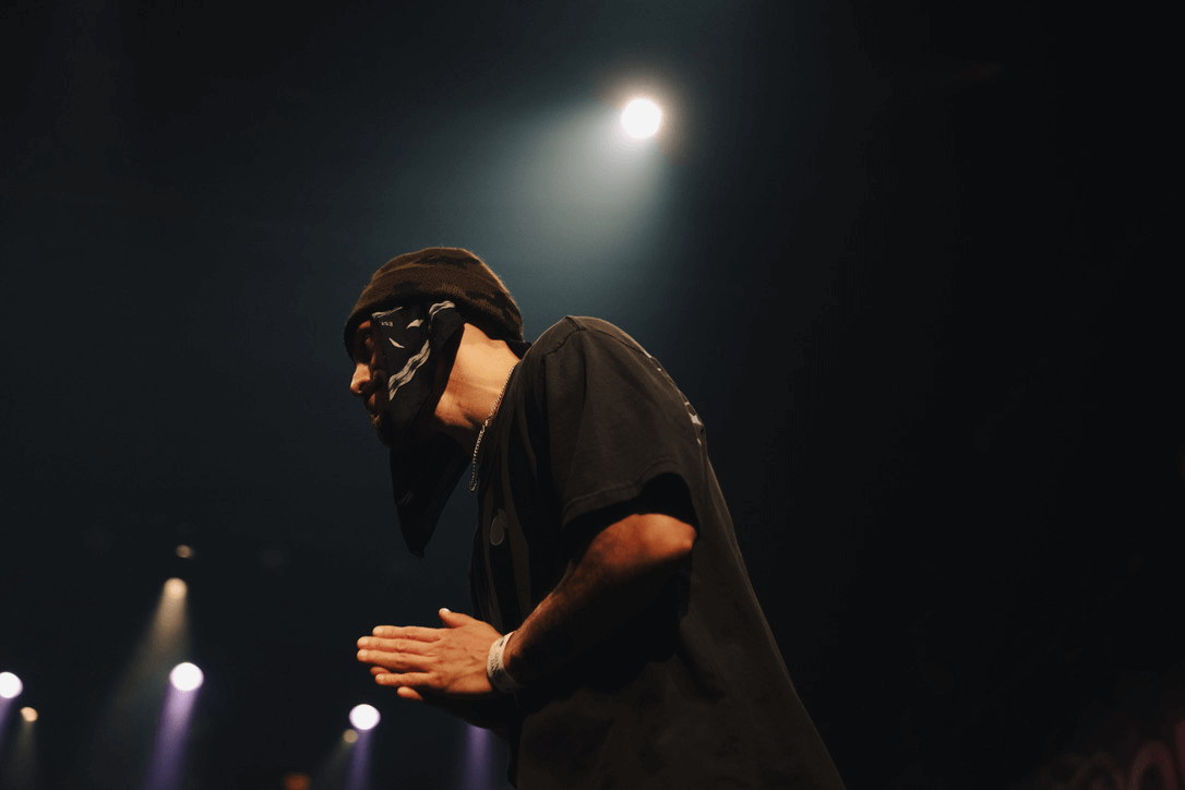 A masked figure in a beanie bowing with hands together under stage light — the site's closing image.
